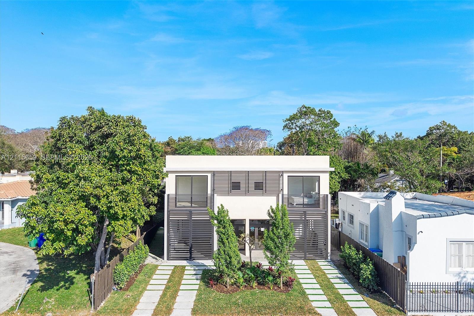 Modern two-story exterior featuring a light-toned stucco facade, horizontal slat privacy screening, double glass balcony doors, and a linear stone-paver driveway