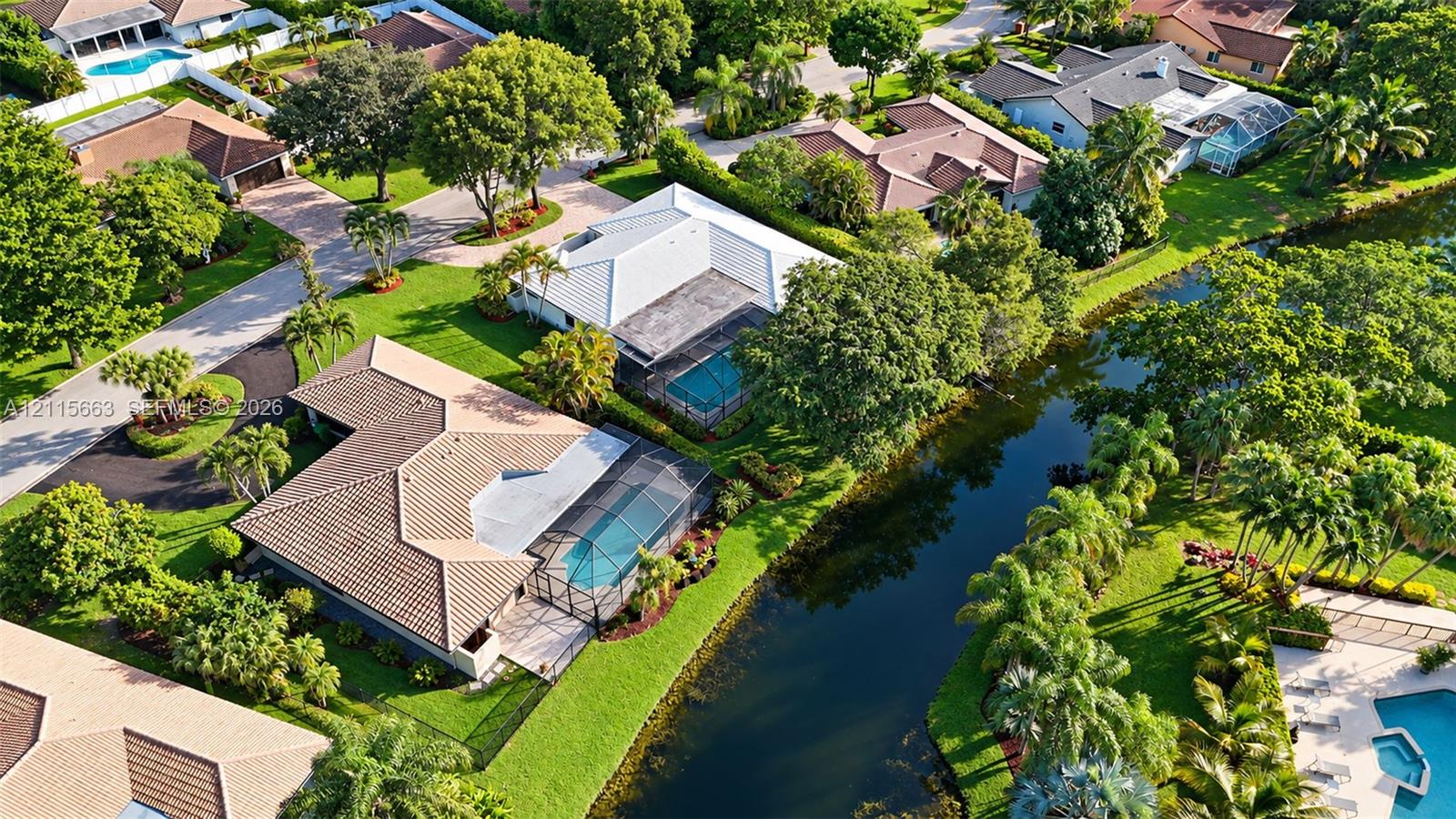 Aerial perspective of a single-story residence featuring a tile roof, an enclosed swimming pool, manicured landscaping, and a canal-front lot