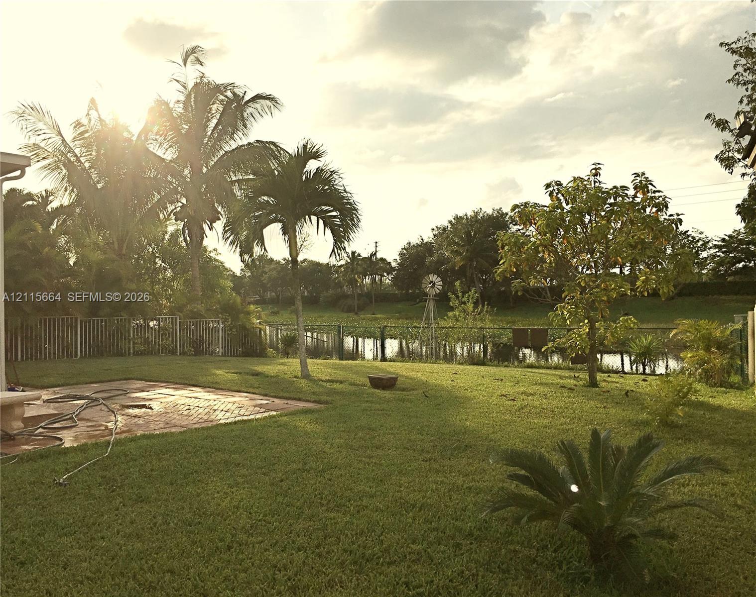 Expansive backyard featuring a brick paver patio, metal perimeter fencing, mature palm trees, and waterfront grounds