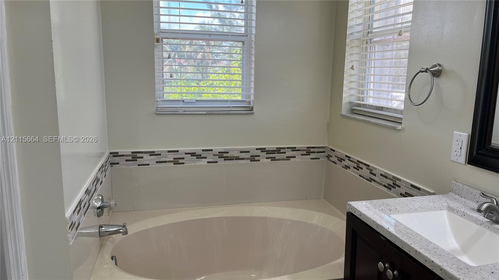 Oval soaking tub with chrome hardware, decorative horizontal tile border, stone-finish countertop, undermount sink, and dark wood-tone cabinetry