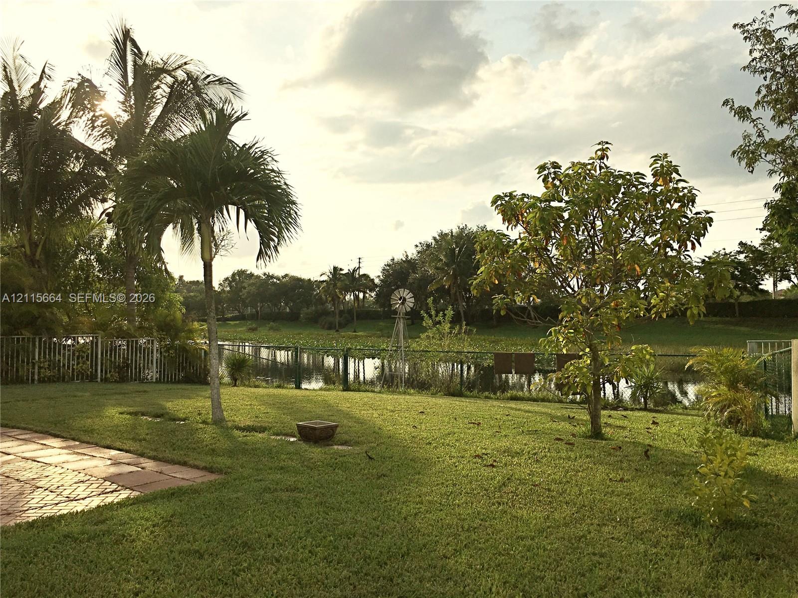 Backyard featuring a brick-paver patio, expansive lawn, mature palm trees, perimeter fencing, and water frontage