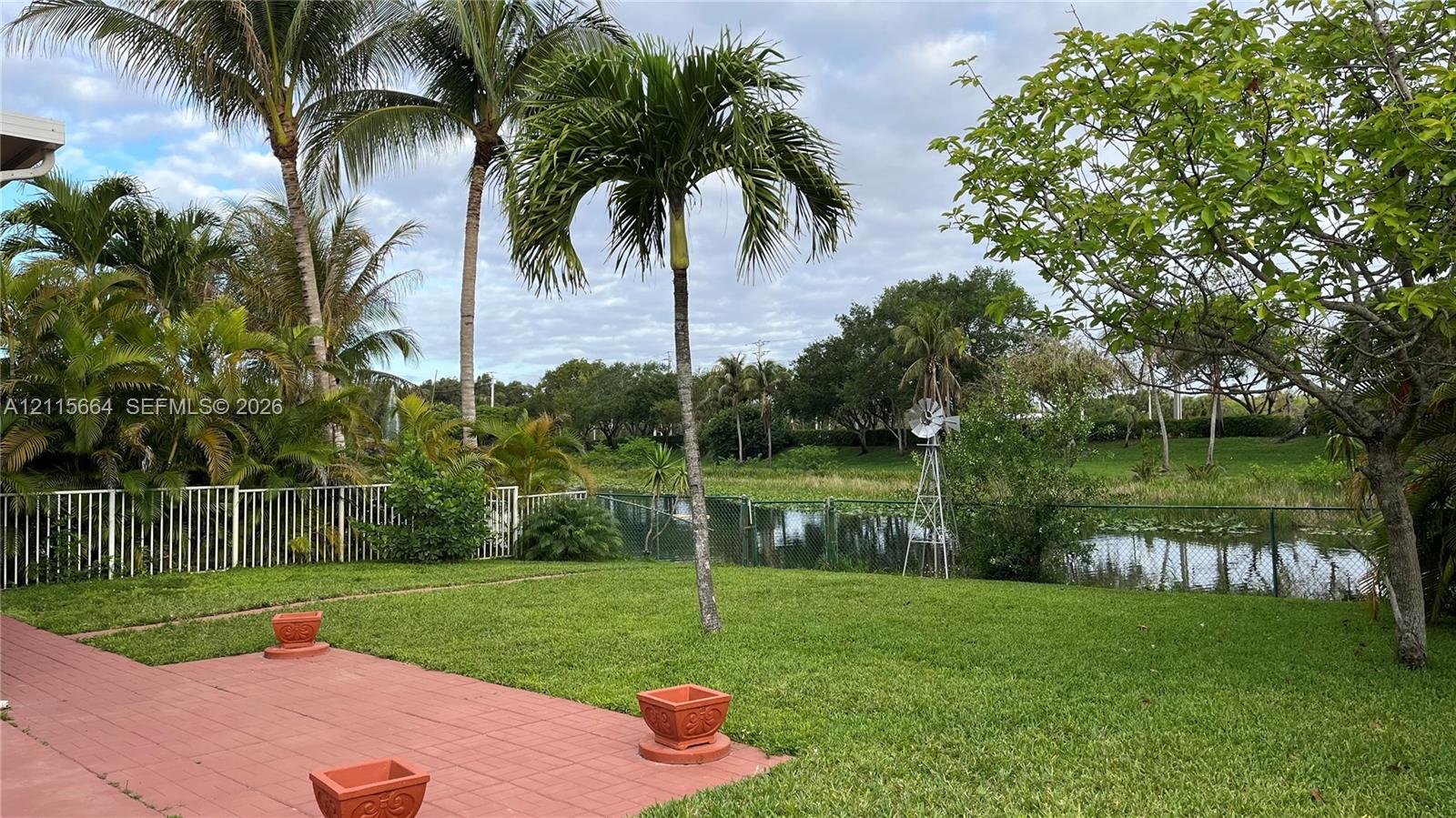 Rear exterior featuring a paved terrace, expansive lawn, mature palm trees, metal fencing, and a waterfront backdrop with a decorative windmill