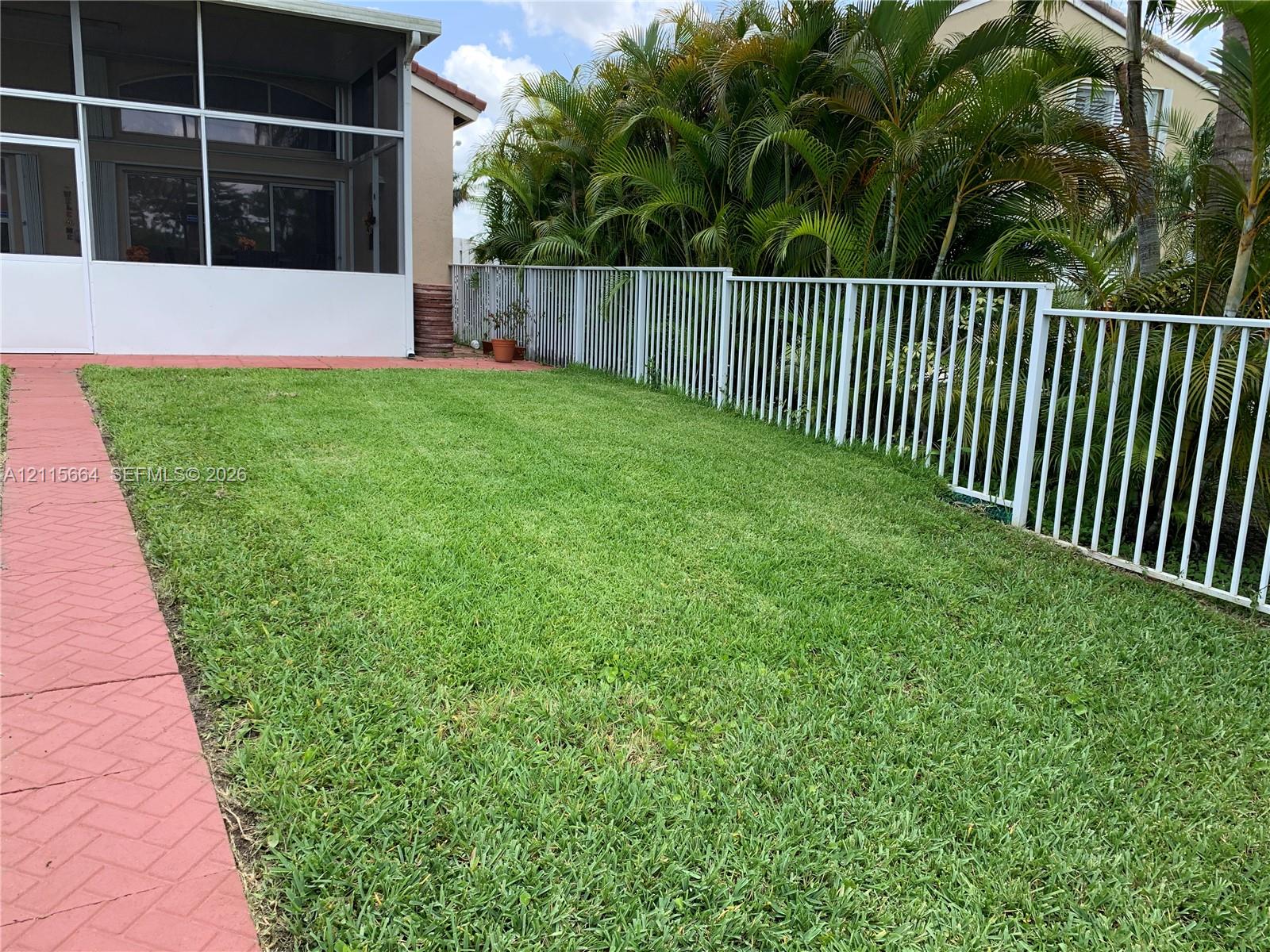 Backyard featuring a grass lawn, white metal fencing, a red brick-pattern paver walkway, and a screened-in porch area