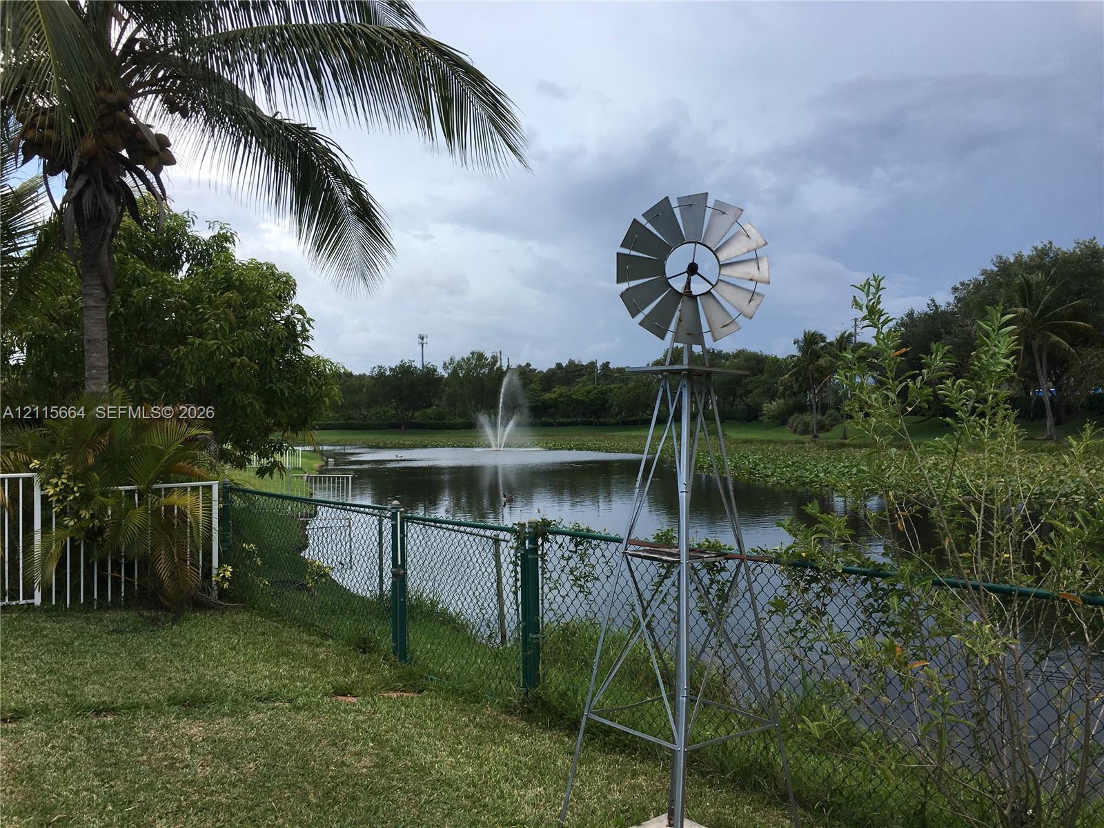 Waterfront grounds featuring a metal windmill, a center-pond fountain, chain-link perimeter fencing, and mature palm trees