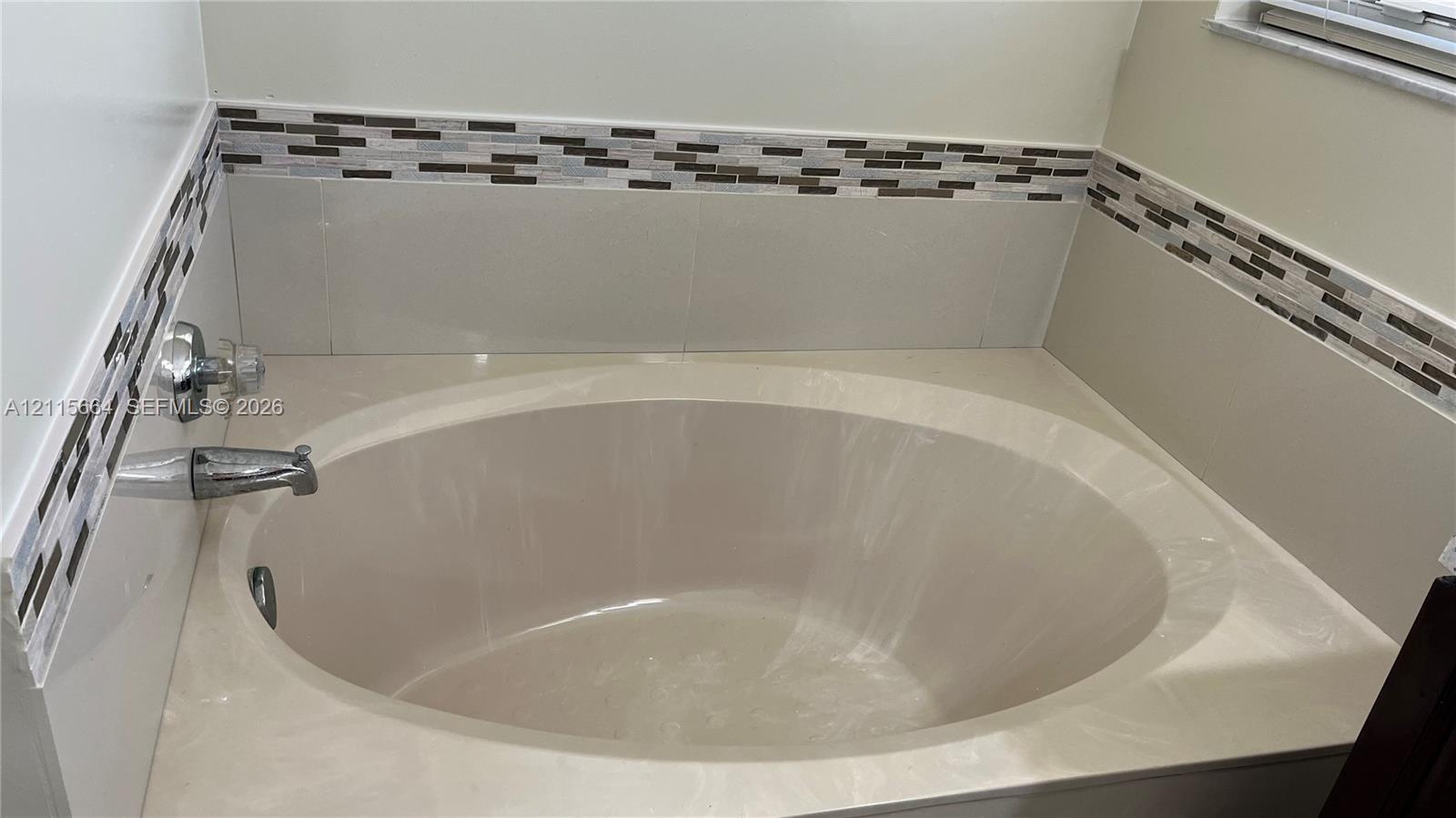 Oval soaking tub featuring an integrated surround, chrome fixtures, and a decorative multi-tone glass tile backsplash