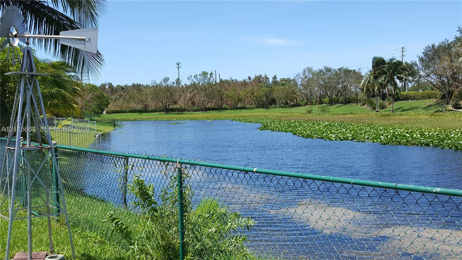 Waterfront lot featuring a metallic windmill, green chain-link perimeter fencing, and mature vegetation