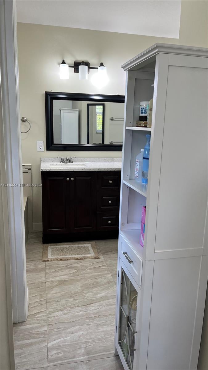 Dark-finish vanity cabinet with a stone-top counter and integrated sink, large framed wall mirror, three-bulb vanity light fixture, and tile flooring
