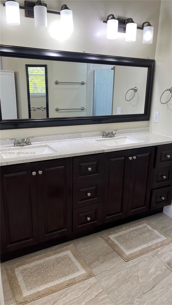 Dual-sink vanity featuring dark wood cabinetry, speckled stone-composite countertops, large framed mirror, and two multi-bulb overhead light fixtures