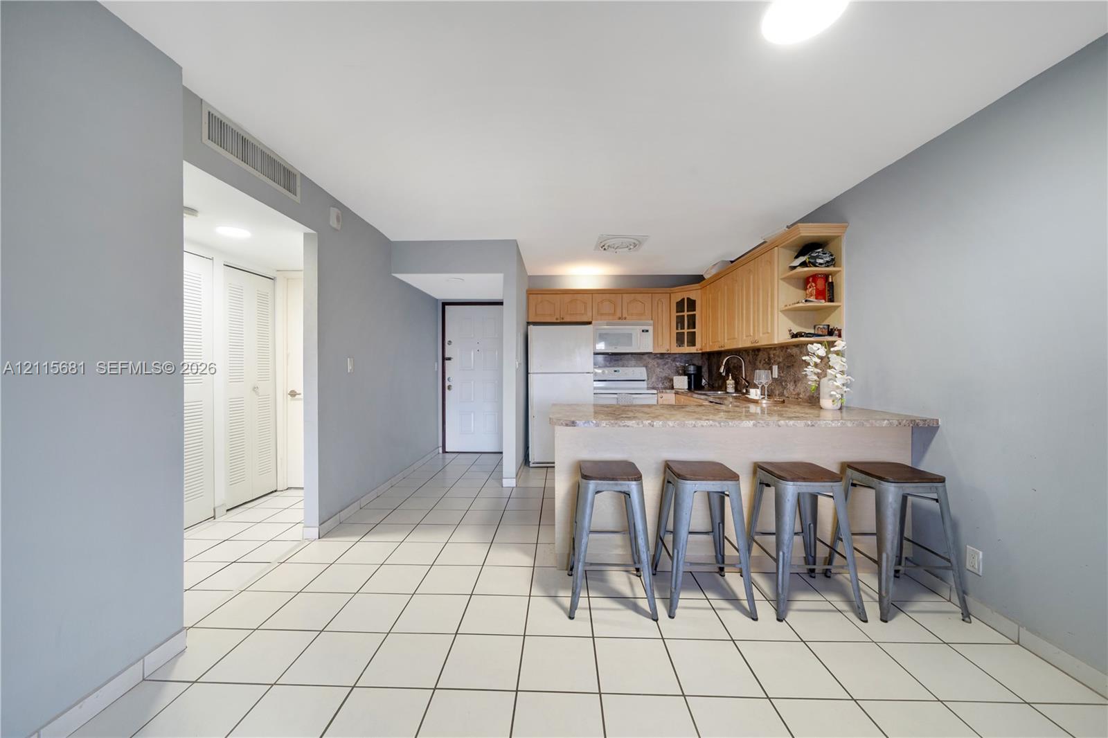 Kitchen area featuring wood-finish cabinetry, stone-patterned countertops, integrated breakfast bar, white appliance suite, and neutral tile flooring