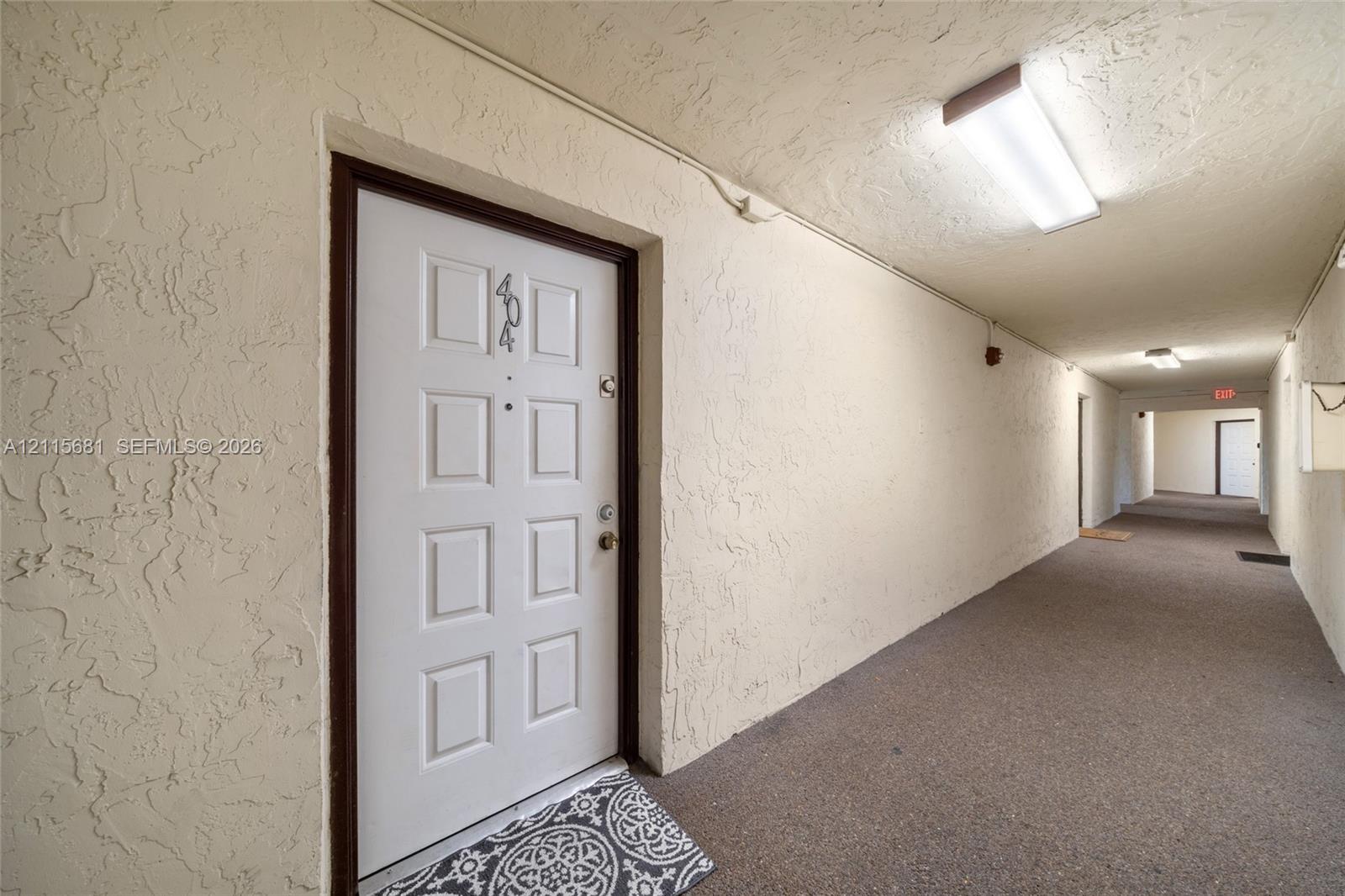 White six-panel entry door with integrated peephole and metallic hardware, textured wall finishes, and overhead surface-mounted lighting