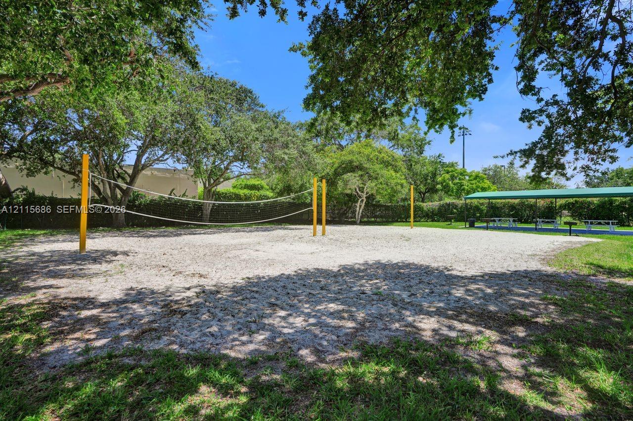 Sand volleyball court featuring a netted boundary, perimeter landscaping, mature shade trees, and a covered pavilion