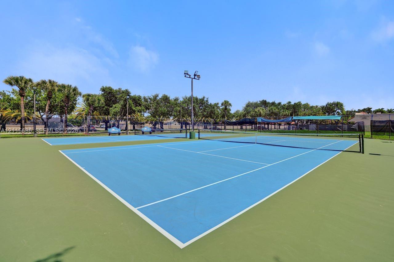 Dual tennis courts featuring a synthetic surface, perimeter chain-link fencing, integrated lighting poles, and mature tree landscaping