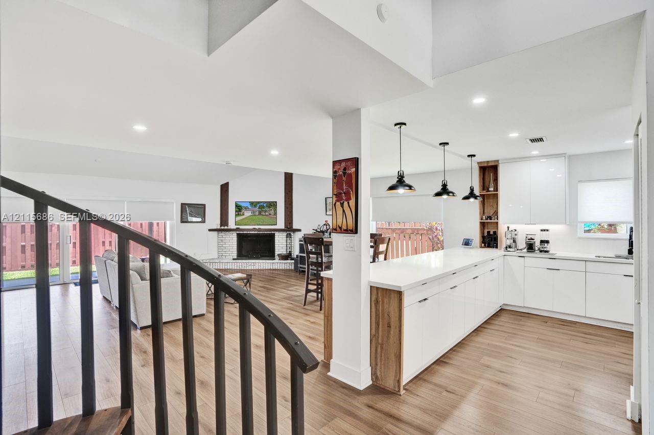 Open-concept kitchen featuring high-gloss white cabinetry, expansive countertop peninsula, wood-finish flooring, and industrial-style pendant lighting, complemented by a floor-to-ceiling brick fireplace