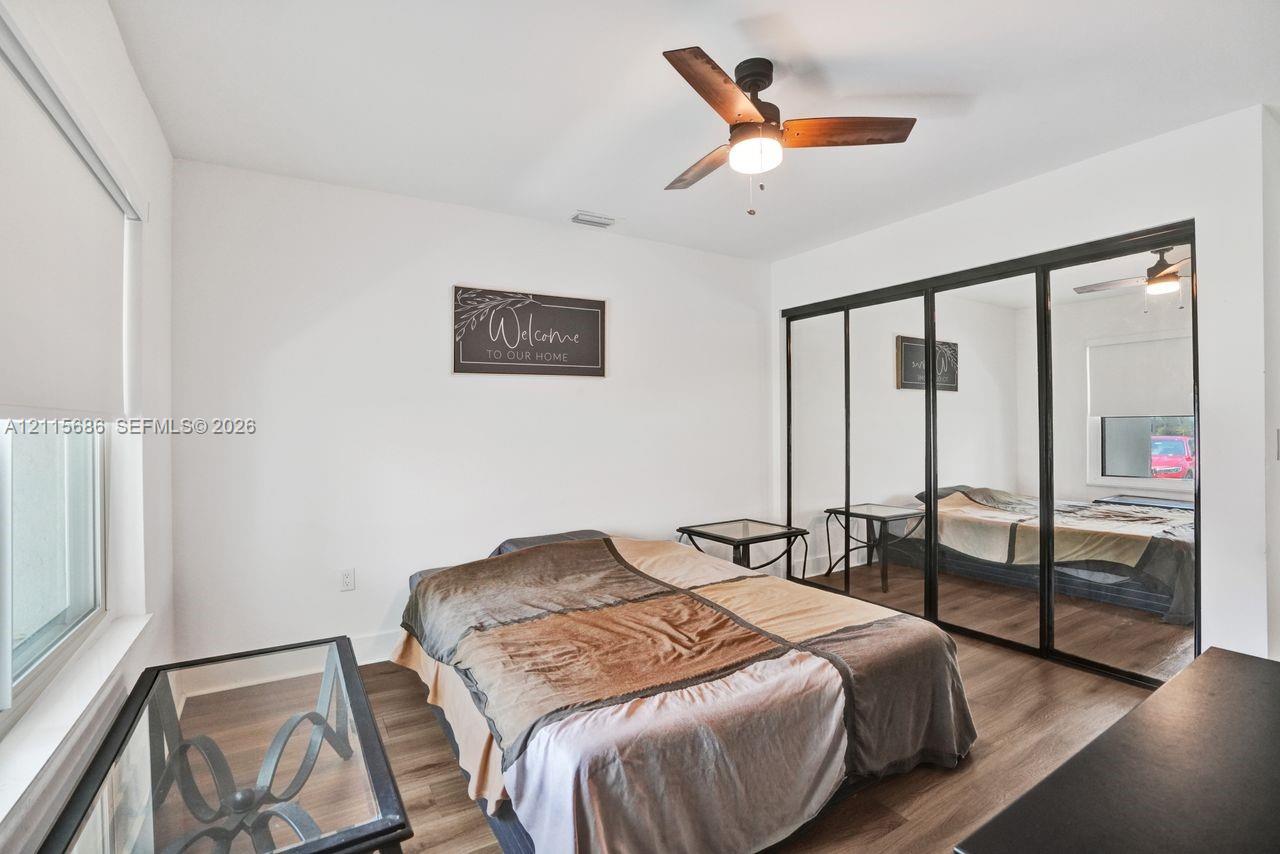 Wood-finish flooring, sliding mirror-paneled closet doors with black frames, modern ceiling fan with integrated lighting, and roller window shades