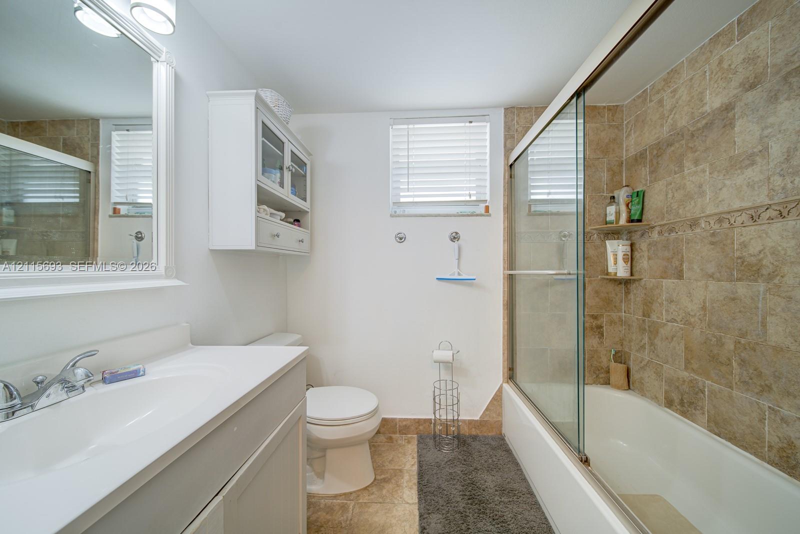 Bathroom featuring a white vanity cabinet with an integrated sink, wall-mounted cabinetry, a glass-enclosed bathtub shower combination, and stone-finish tile surround
