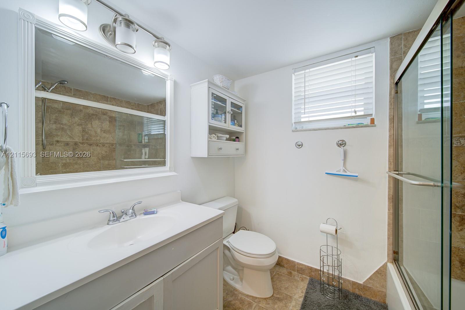 Bathroom featuring a vanity with a white countertop, wall-mounted storage cabinetry, a framed vanity mirror with tri-light fixture, tiled shower surround with glass sliding doors, and tiled flooring