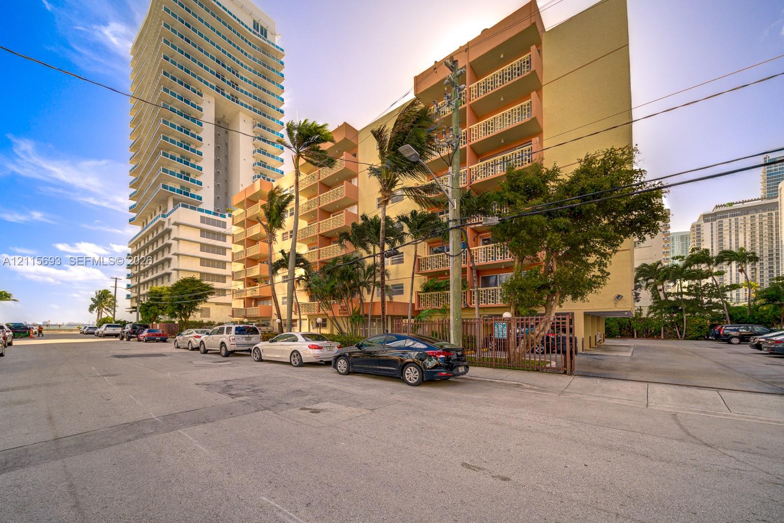 Multi-story residential building featuring private balconies with decorative railings, ground-level surface parking, and mature palm landscaping