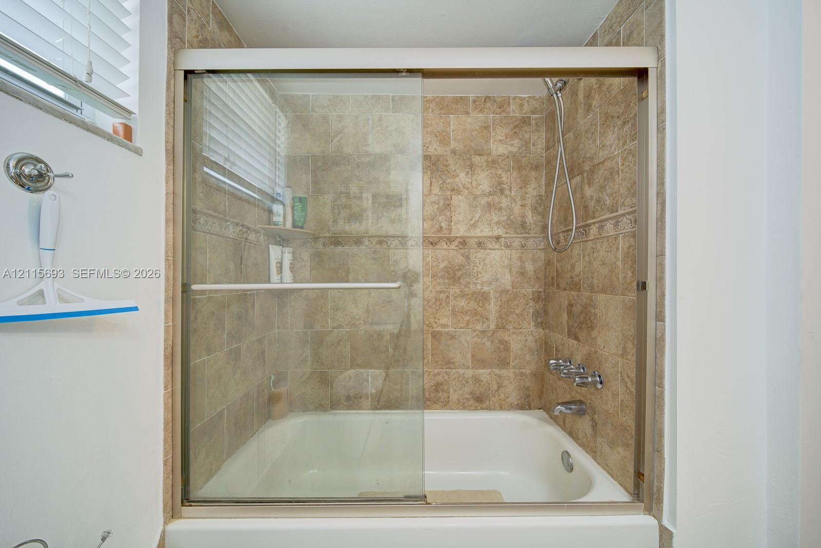 Shower and bathtub combination featuring a sliding glass enclosure, floor-to-ceiling textured tile surround, wall-mounted handheld showerhead, and integrated soap niche