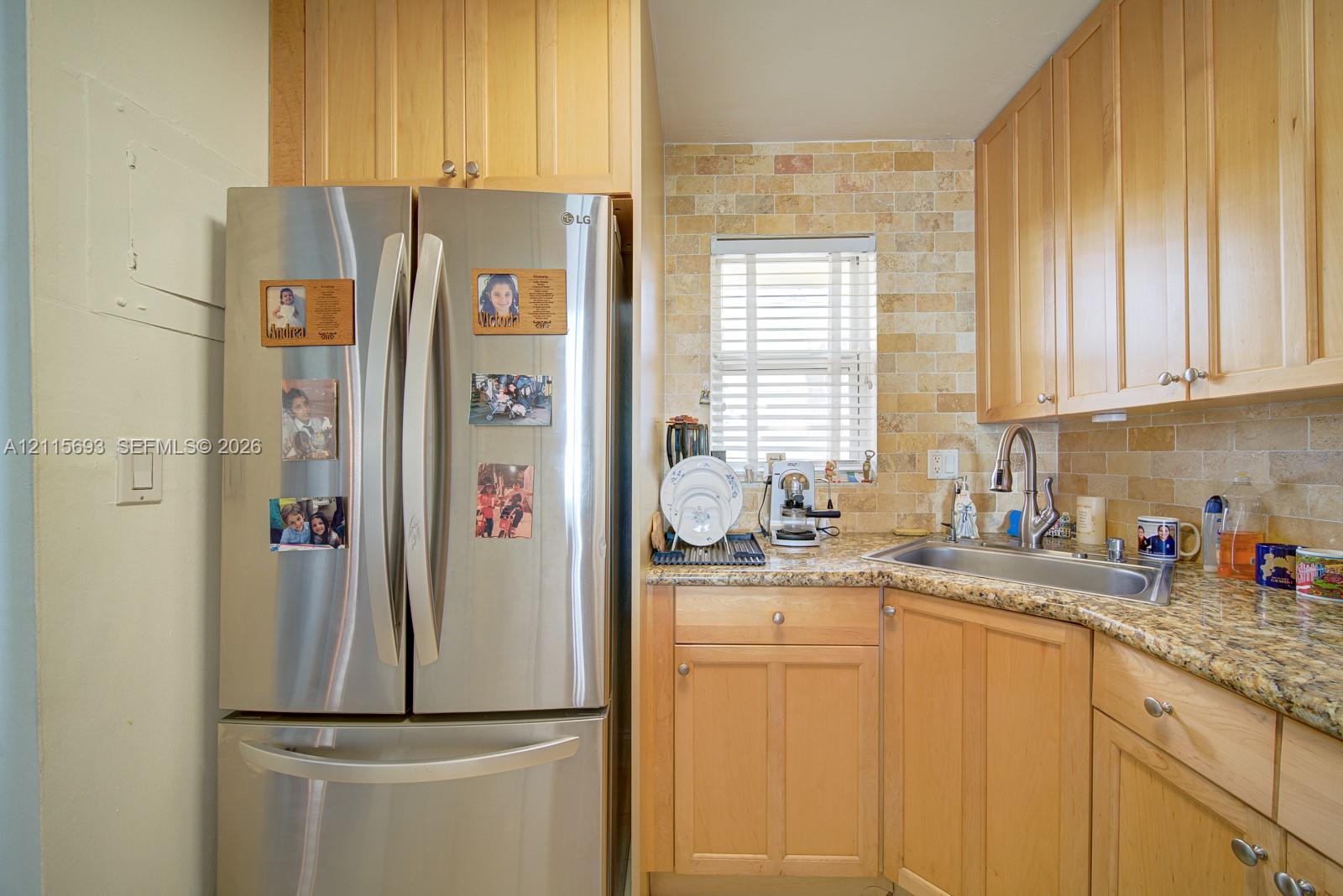 Kitchen featuring light wood cabinetry, stainless steel French door refrigerator, natural stone countertop, tile backsplash, and undermount sink with high-arc faucet