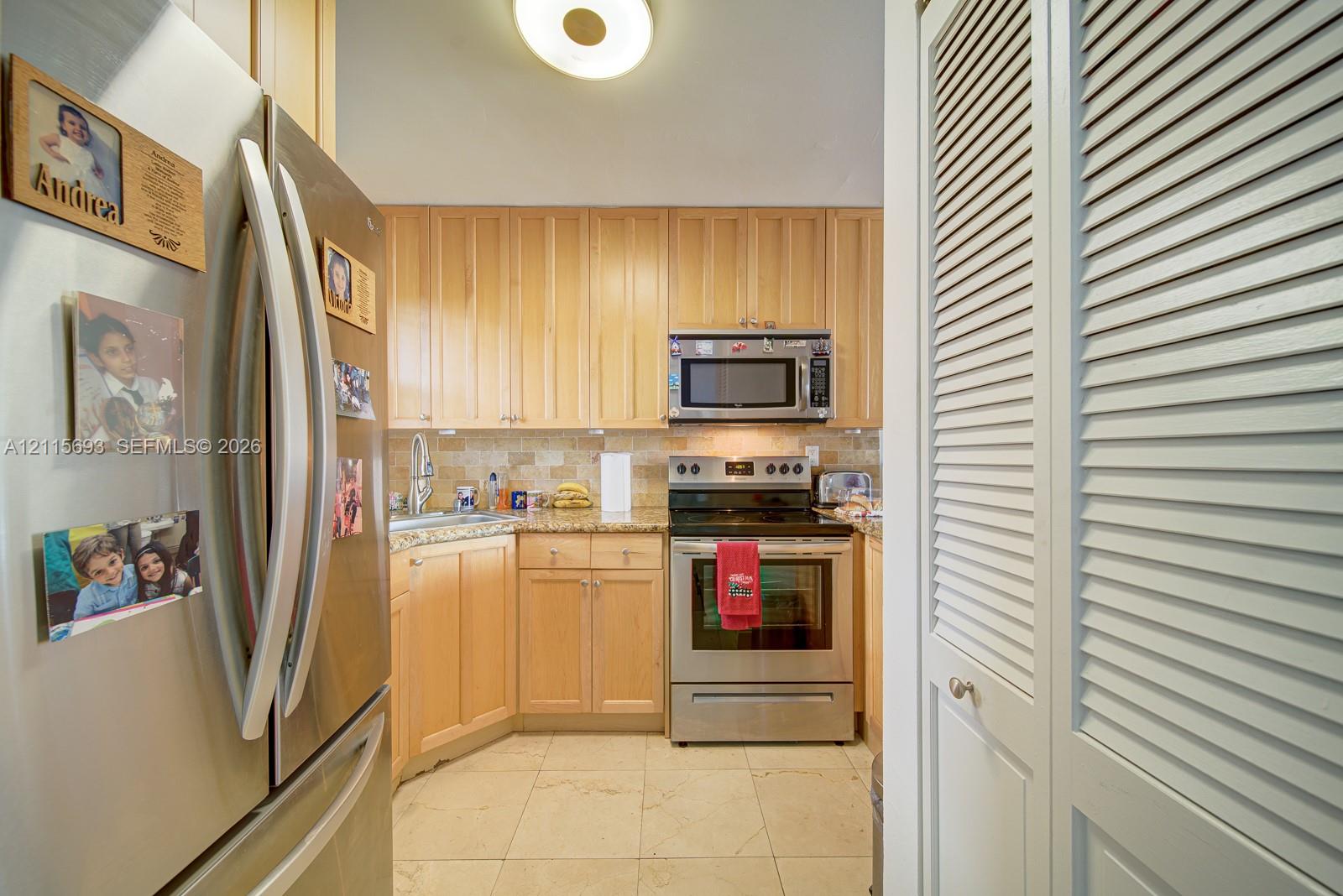 Kitchen featuring light wood cabinetry, stone countertops, tiled backsplash, stainless steel appliances, and louvered closet doors