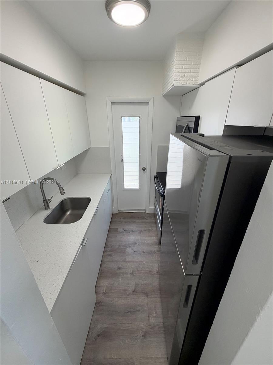Galley kitchen featuring flat-panel white cabinetry, solid surface countertops, stainless steel sink with pull-down faucet, wood-finish flooring, and a glass-paneled exterior door