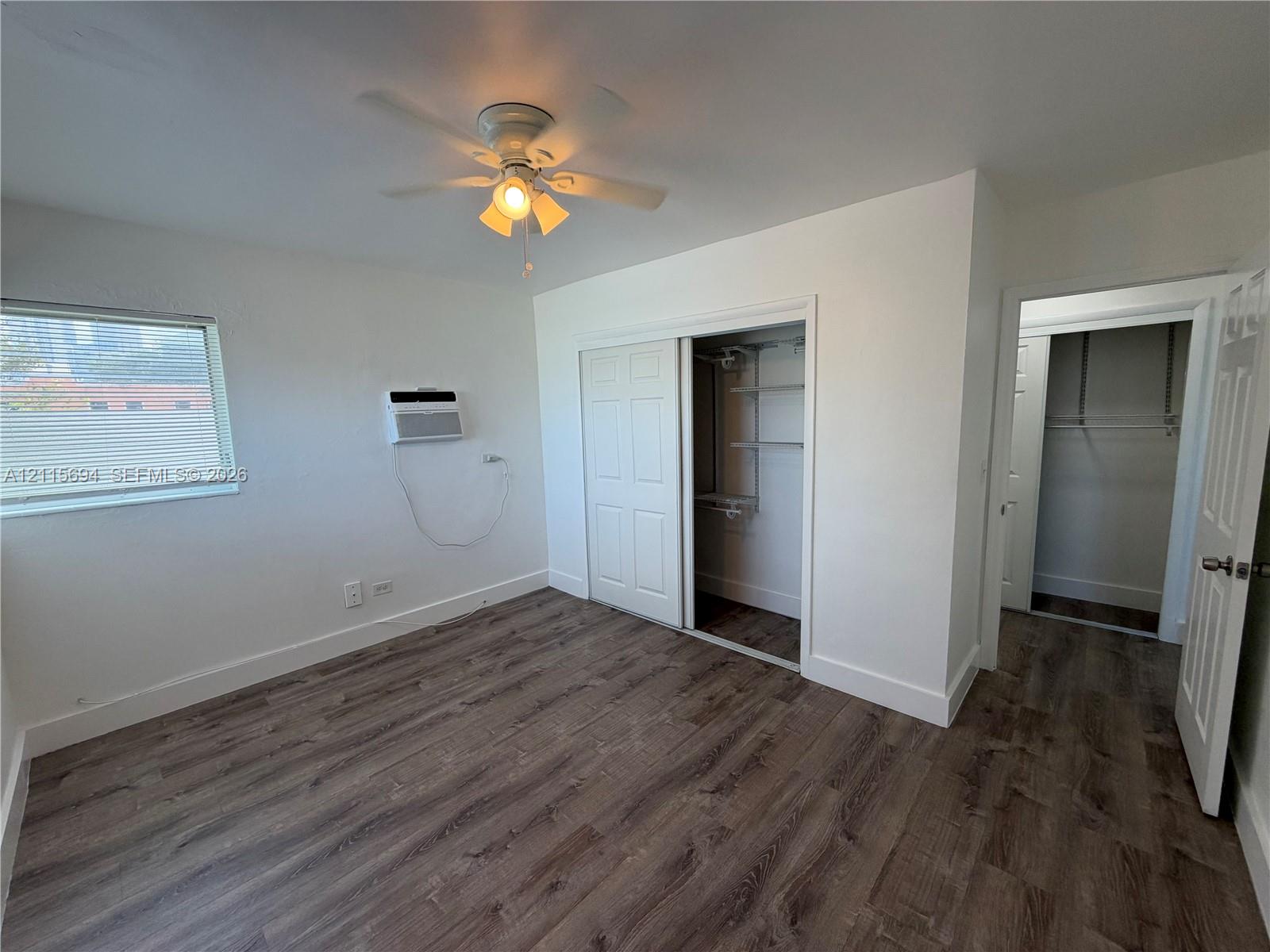 Wood-finish flooring, ceiling fan with integrated lighting, window with horizontal blinds, built-in closet storage, and wall-mounted air conditioning unit