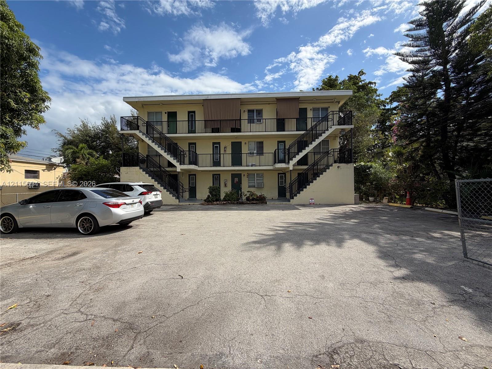 Three-story exterior featuring a light-toned stucco faade, metal balcony railings, external staircases, and an asphalt-paved surface parking lot Three-story exterior featuring a light-toned stucco faade, metal balcony railings, external staircases, and an asphalt-paved surface parking lot
