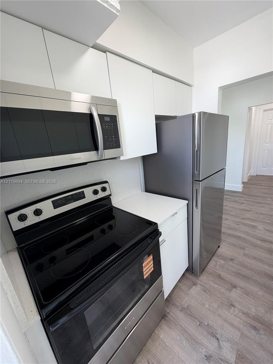 Kitchen featuring white flat-panel cabinetry, stainless steel microwave, glass-top range, stainless steel refrigerator, and wood-finish flooring