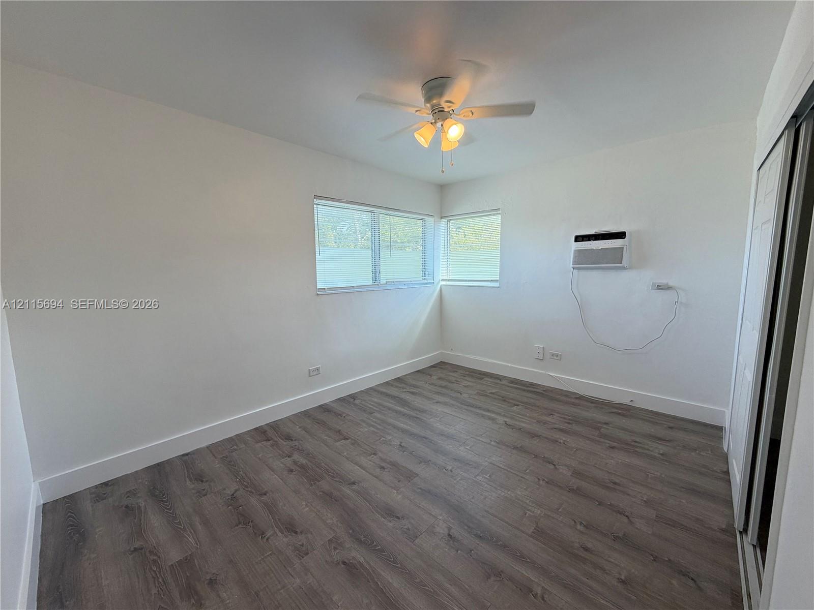 Interior space featuring wood-finish flooring, a ceiling fan with integrated lighting, dual windows, and a wall-mounted air conditioning unit