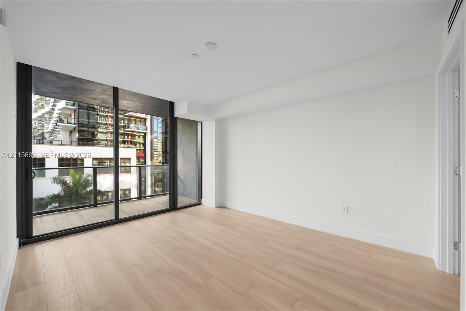 Floor-to-ceiling sliding glass doors, neutral wood-finish flooring, and an attached balcony with black metal railings