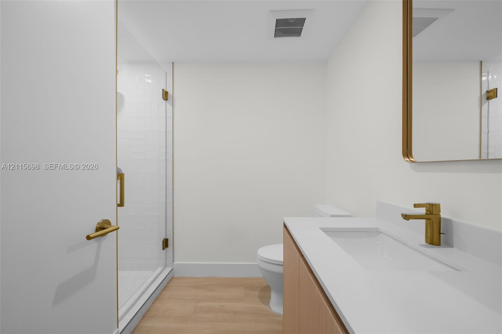 Guest bathroom featuring a walk-in shower with a glass enclosure, wood-finish flooring, vanity with a white countertop and integrated sink, brass-toned hardware, and a matching framed mirror