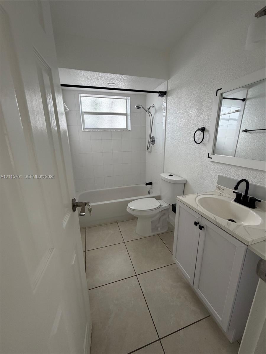 Bathroom featuring large-format floor tile, vanity with stone-surfaced countertop, integrated sink with matte black faucet, and a shower-tub combination with white tile surround