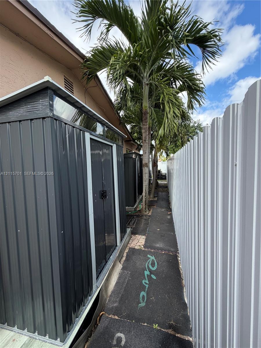 Metal storage structure featuring a gable roof, corrugated siding, and double doors, situated along a paved exterior pathway bordered by a metal privacy fence