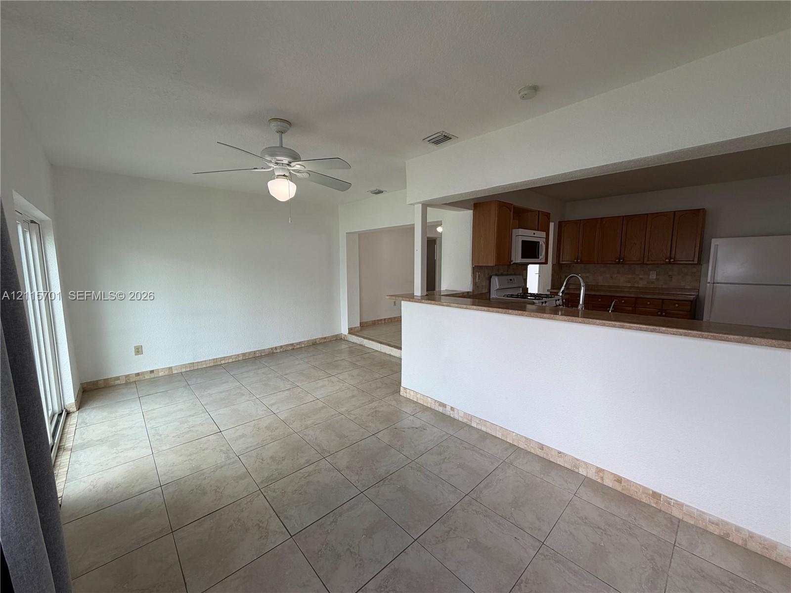 Open-concept interior featuring large-format ceramic tile flooring, a breakfast bar with stone-finish countertops, warm-toned wood cabinetry, and a ceiling fan with integrated lighting Open-concept interior featuring large-format ceramic tile flooring, a breakfast bar with stone-finish countertops, warm-toned wood cabinetry, and a ceiling fan with integrated lighting