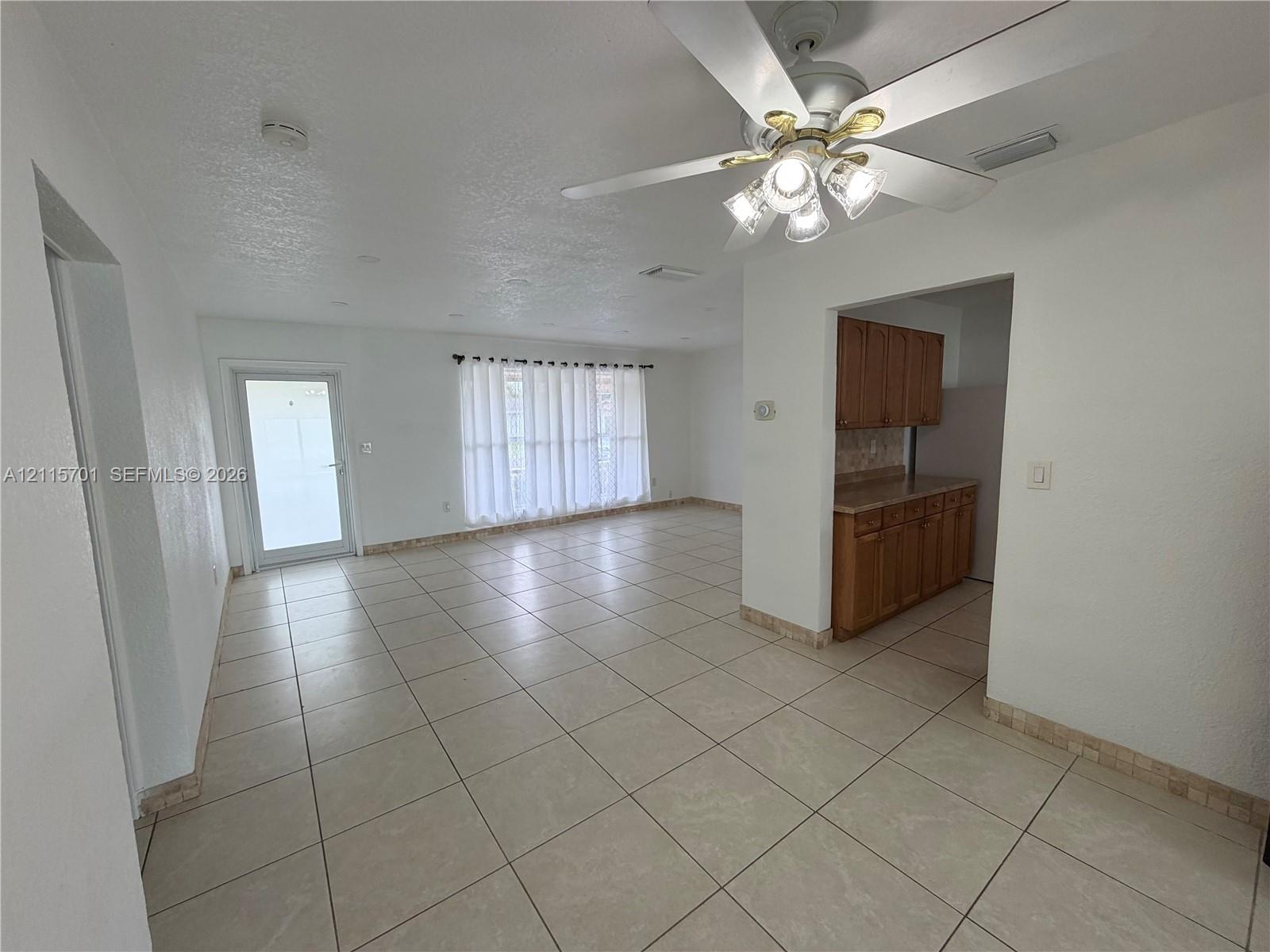 Open interior space featuring light-toned tile flooring, white-finish walls, ceiling-mounted paddle fan with integrated light fixture, and wood-finish cabinetry Open interior space featuring light-toned tile flooring, white-finish walls, ceiling-mounted paddle fan with integrated light fixture, and wood-finish cabinetry