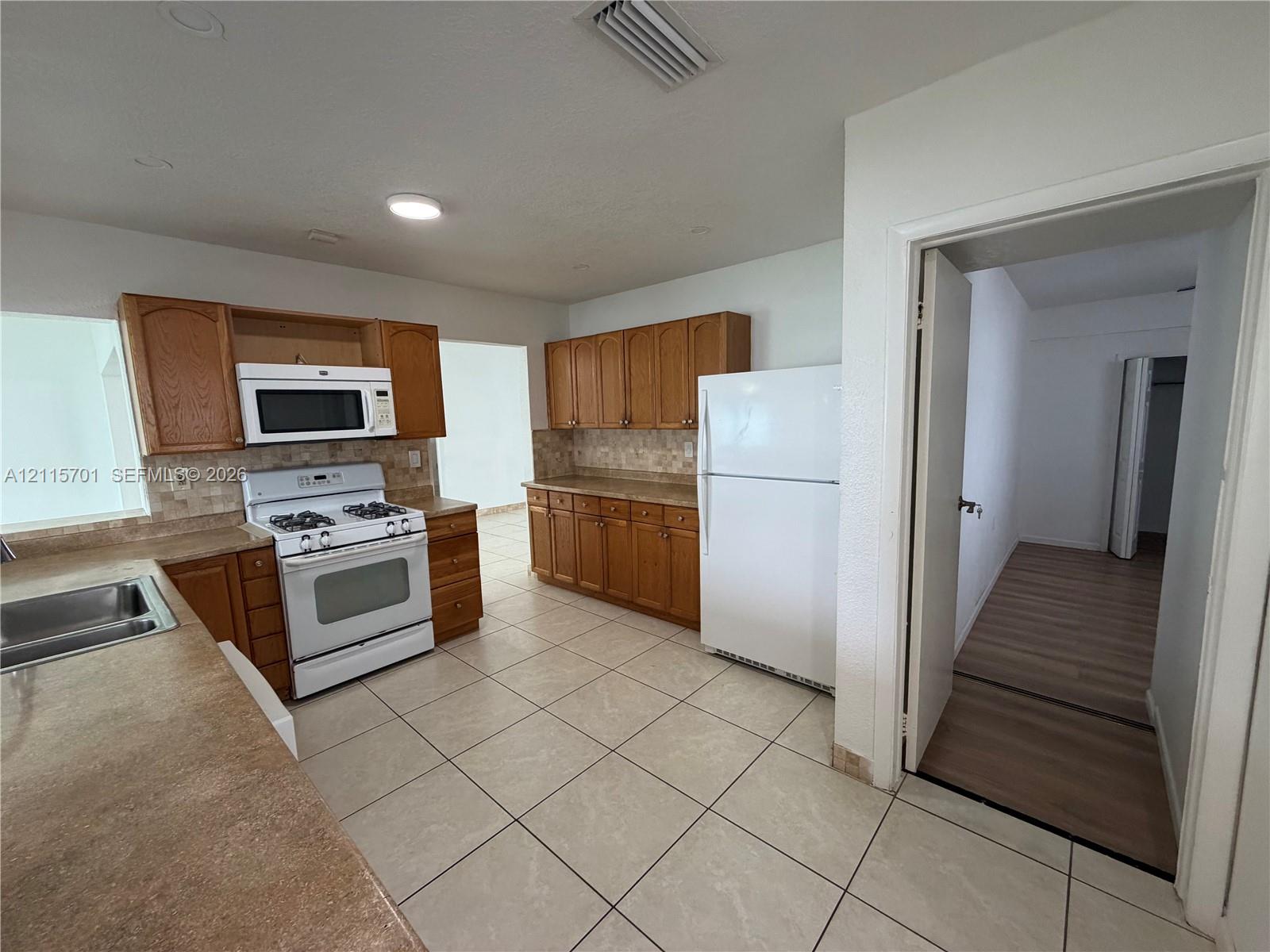 Kitchen featuring wood cabinetry, tile flooring, stone-finish countertops, tile backsplash, and white appliance suite