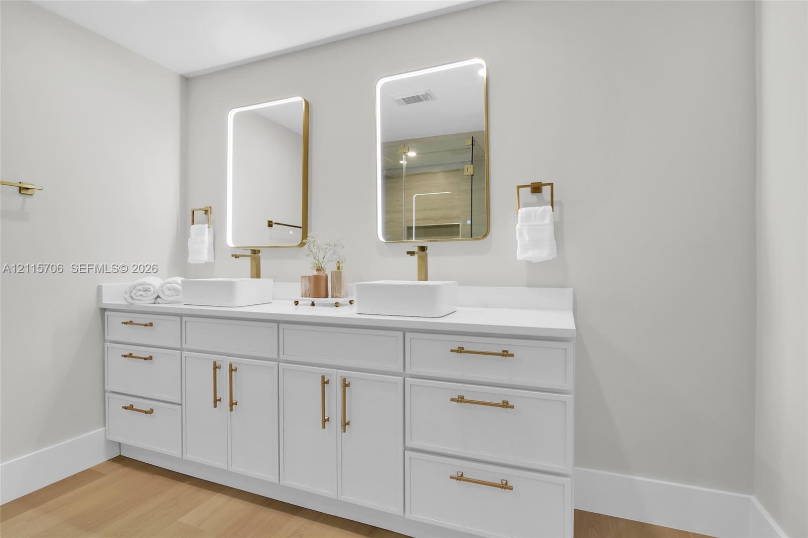 Double vanity with white cabinetry, gold-tone hardware, rectangular vessel sinks, quartz countertops, and lighted mirrors with gold-tone frames