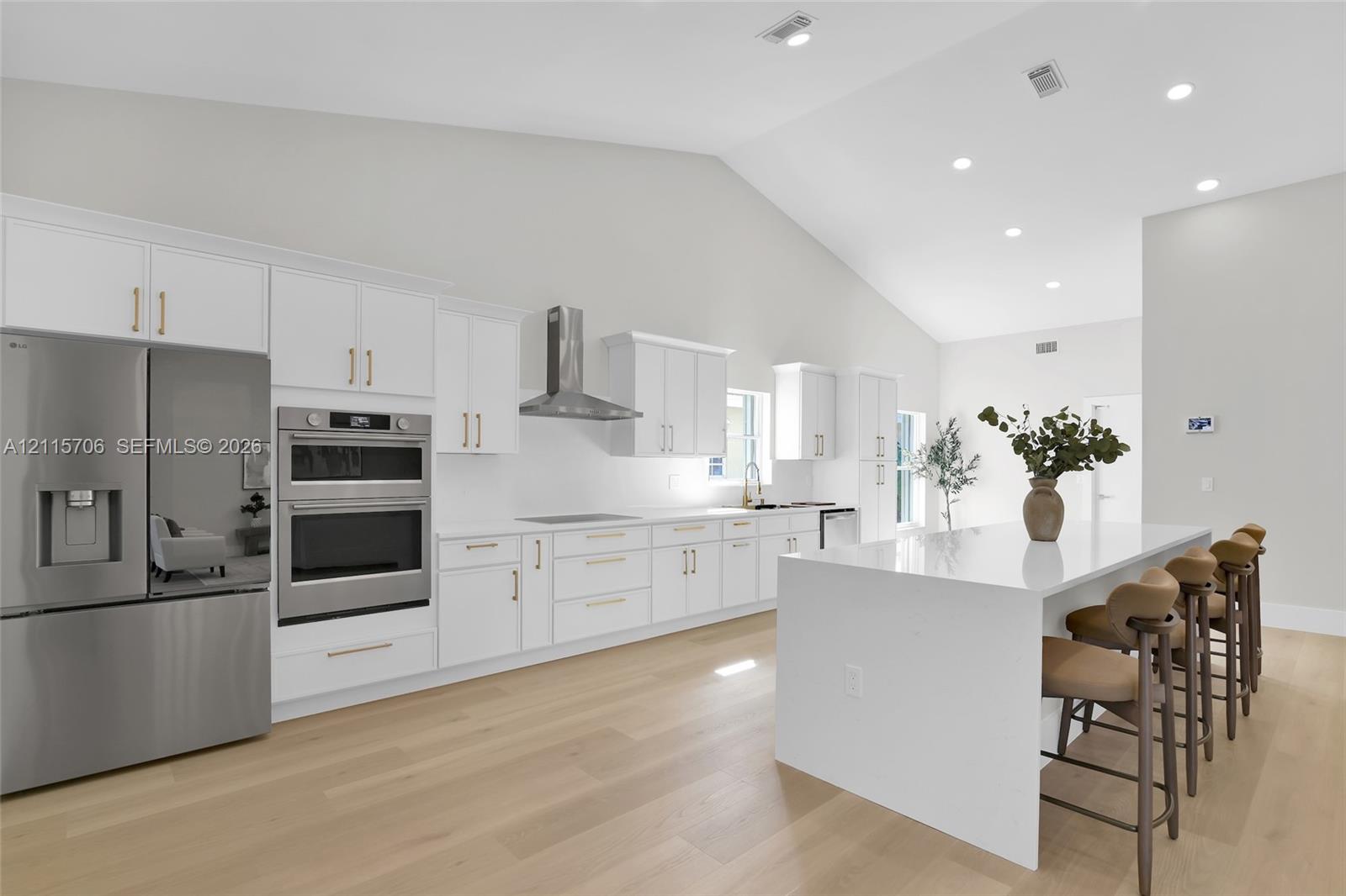 Kitchen featuring vaulted ceilings, white shaker-style cabinetry with gold-tone hardware, stainless steel built-in double oven, matching French-door refrigerator, and a waterfall-edge center island with stone-finish countertop