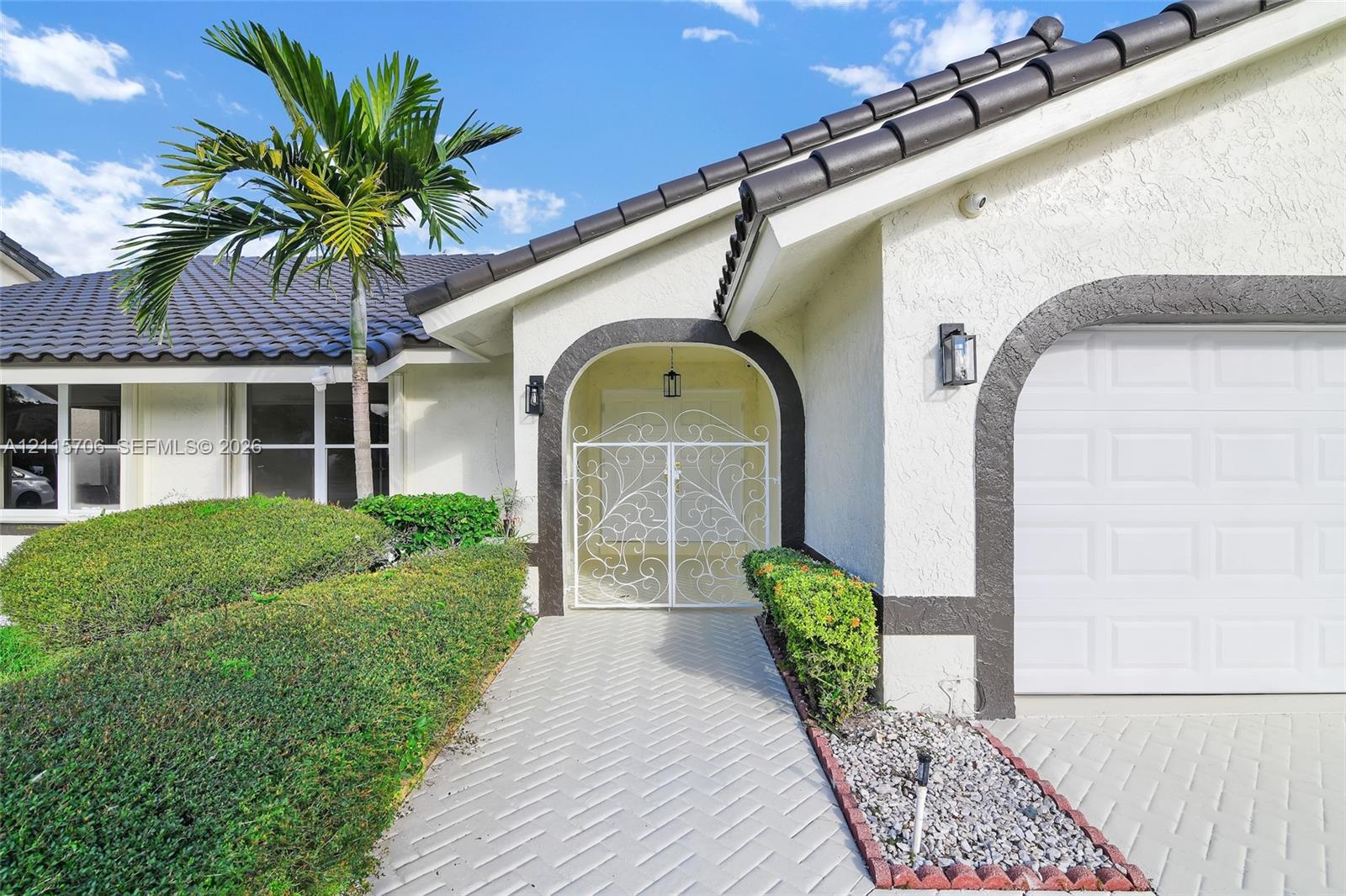 Stucco exterior with a tiled roof, arched entryway featuring a decorative metal gate, herringbone-patterned paver walkway, and manicured shrubbery