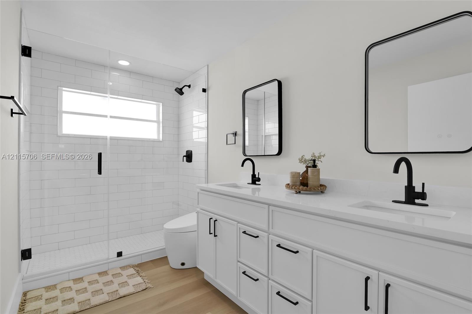 Dual-vanity cabinetry with white surfaces and integrated rectangular sinks, accented by matte black fixtures, framed mirrors, a walk-in shower with glass enclosure, and wood-finish flooring