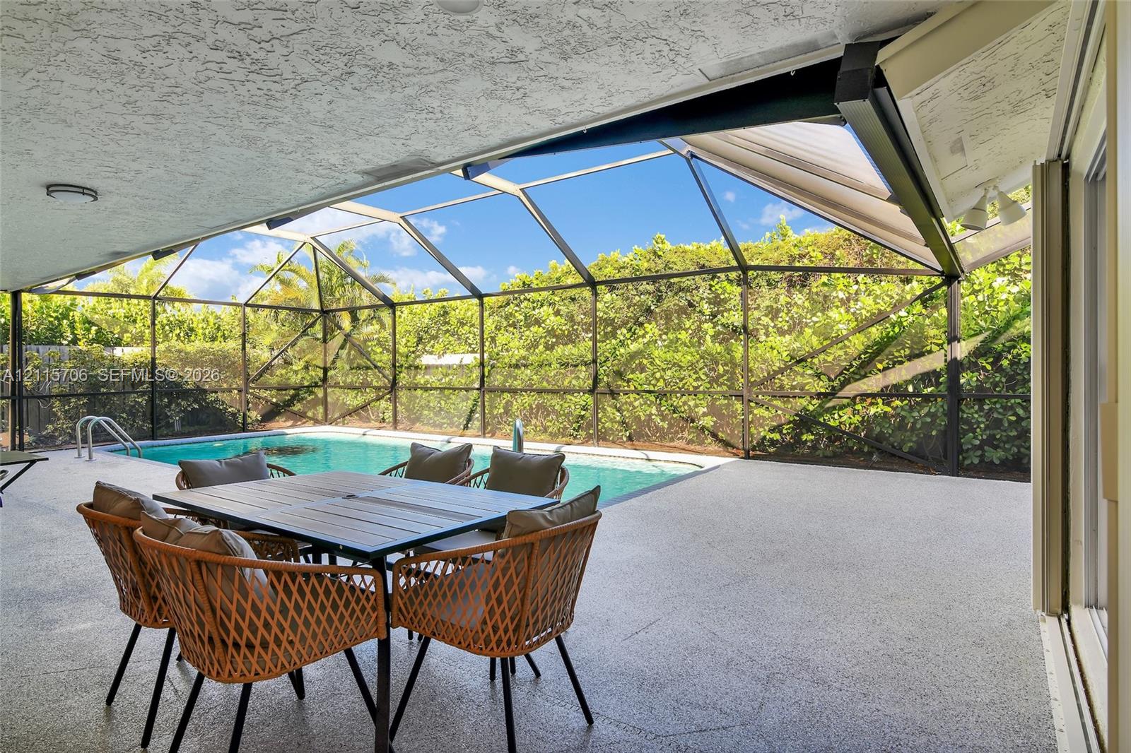 Covered lanai with textured concrete flooring, enclosed swimming pool with metal screen framing, and perimeter privacy hedging