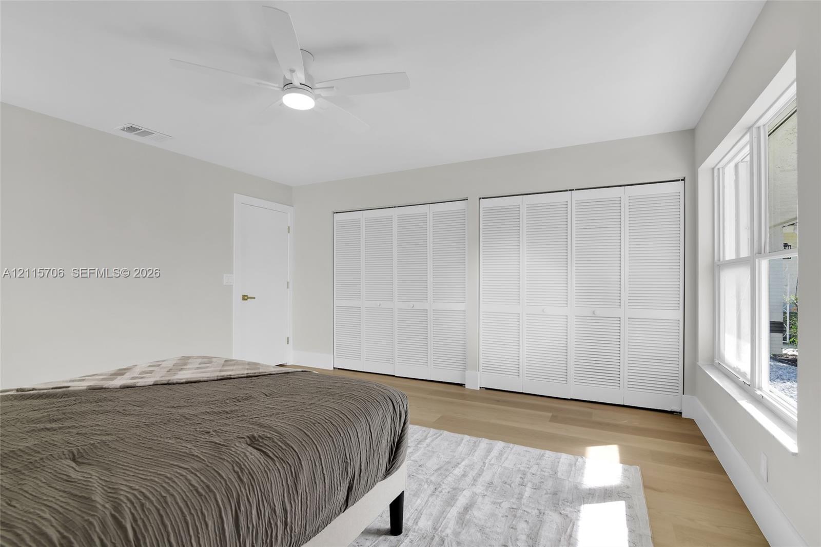 Light-filled room featuring wood-finish flooring, dual sets of white louvered-style closet doors, and a ceiling-mounted fan with integrated lighting