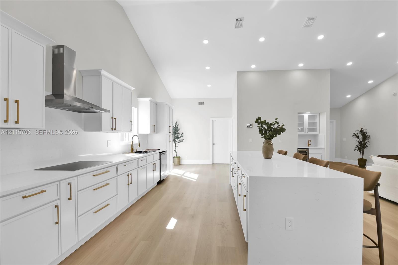 Kitchen featuring shaker-style cabinetry with gold-toned hardware, quartz countertops, a waterfall-edge center island, stainless steel range hood, and wood-finish flooring under vaulted ceilings