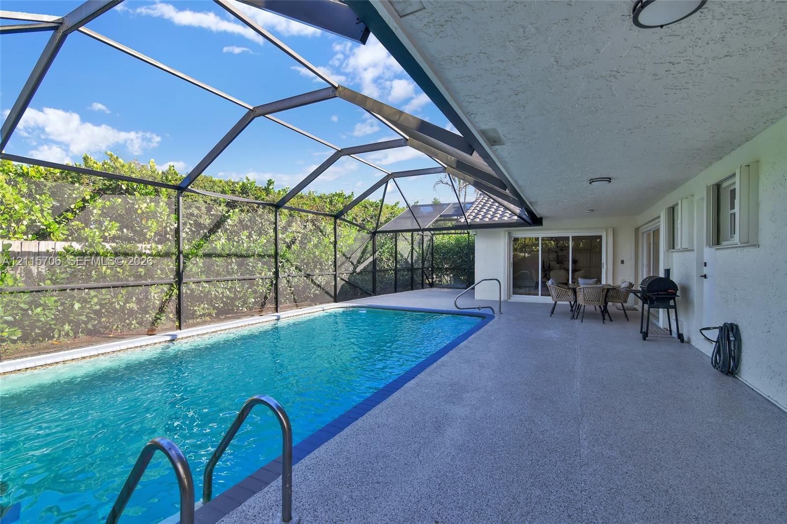 Screened-in outdoor living area featuring a swimming pool with metal ladder, expansive concrete pool deck, and perimeter greenery