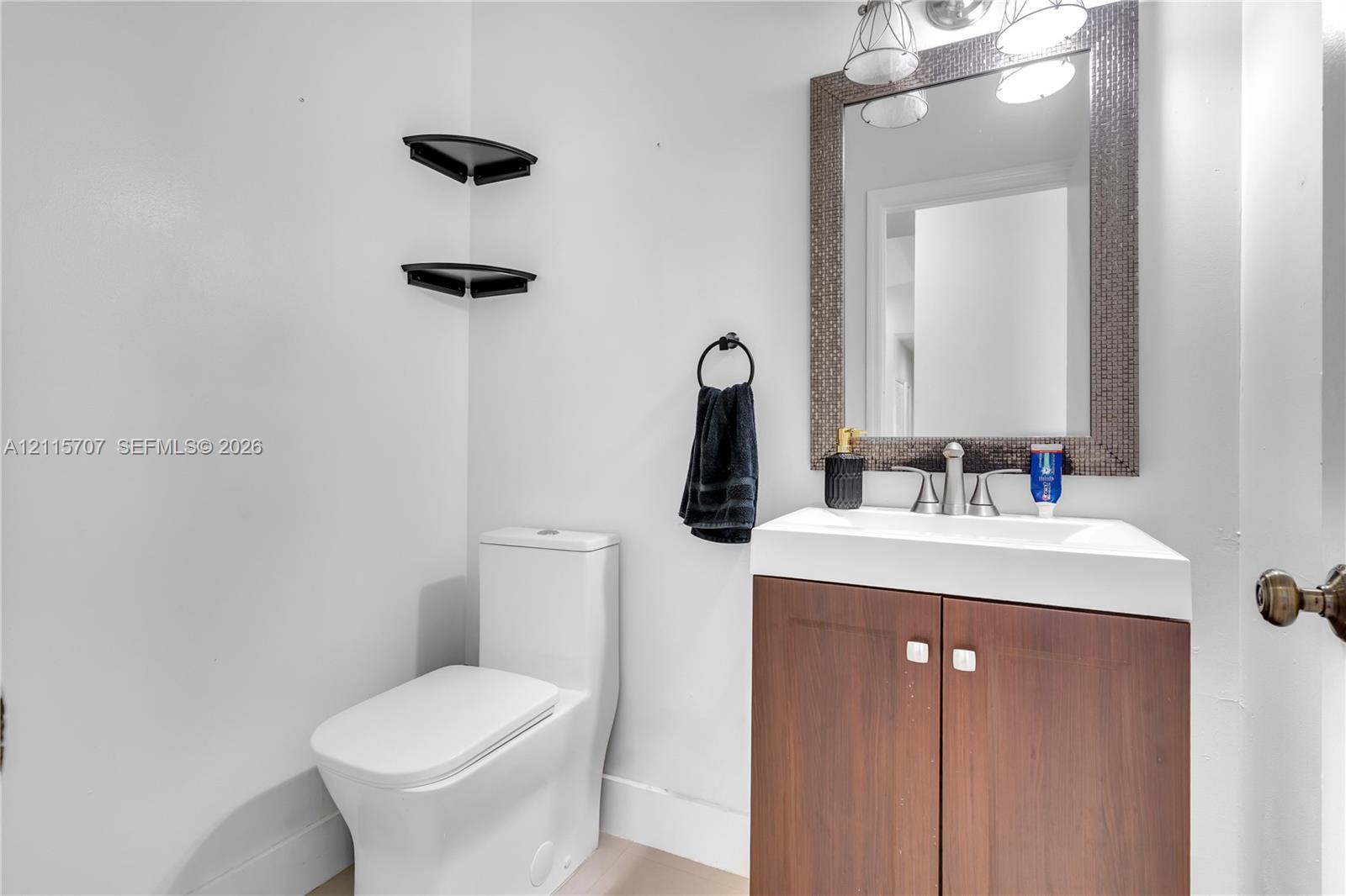Bathroom featuring a wood-finish vanity with a rectangular sink, wall-mounted corner shelving, a framed mirror with integrated lighting, and a modern dual-flush toilet