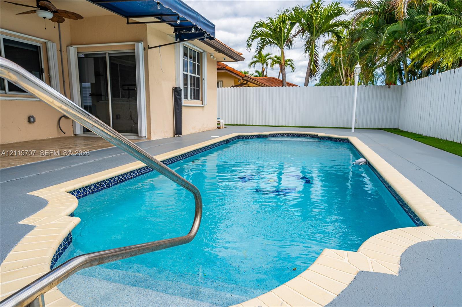 In-ground swimming pool with tile border and integrated steps, concrete pool deck, retractable fabric awning, and white privacy fencing