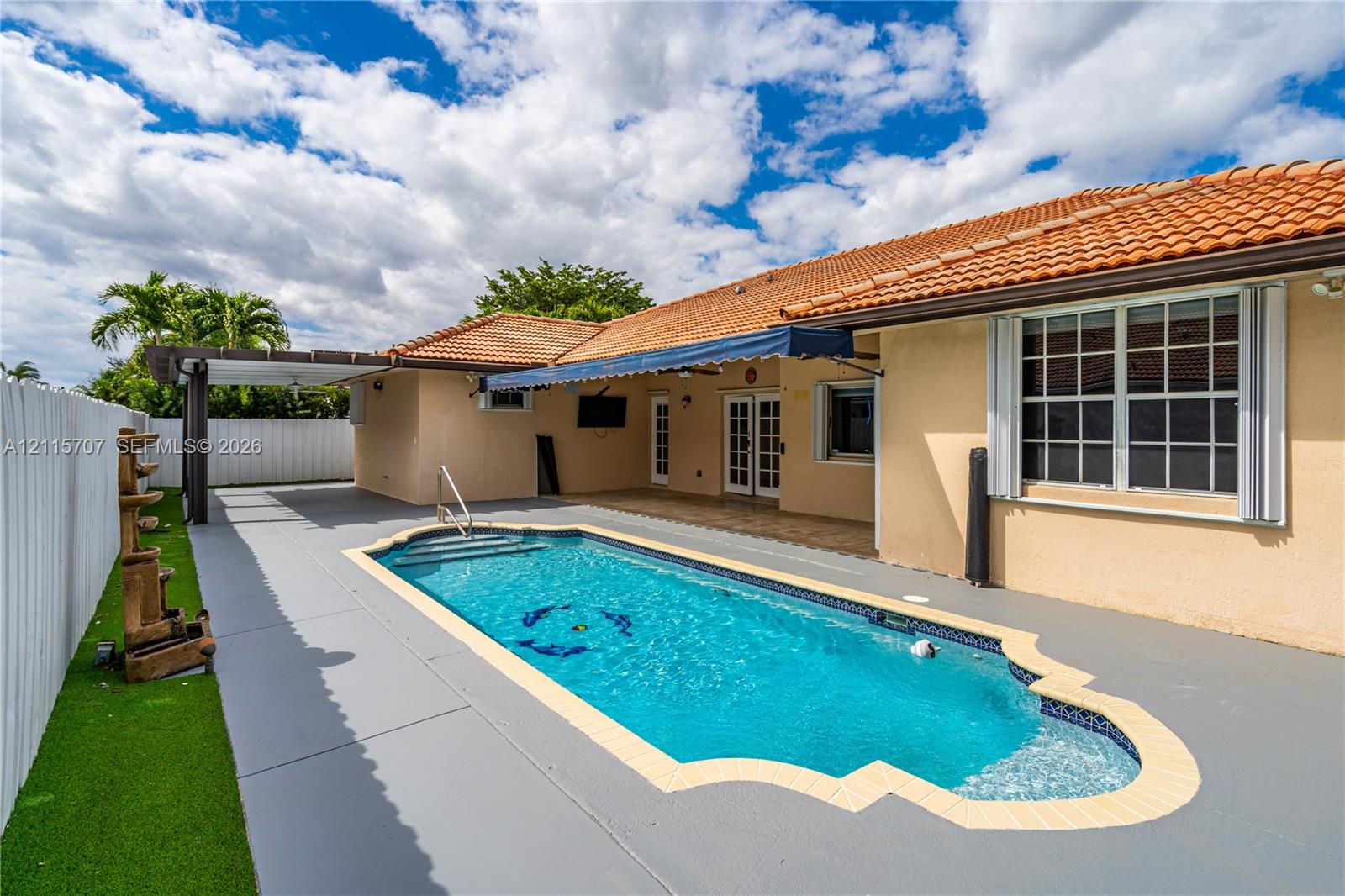 In-ground swimming pool with a custom-shaped perimeter, terra cotta tile roof, retractable fabric awning, wooden-frame pergola, and synthetic turf lawn