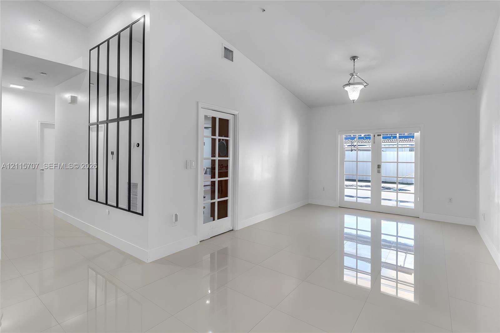 Vaulted ceiling, neutral large-format tile flooring, decorative grid-pattern wall mirror, glass-paned interior door, and double glass-paned exterior doors