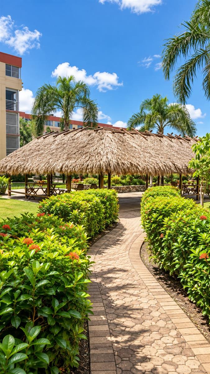 Outdoor pavilion with a thatched roof, brick-paver pathway, and manicured landscaping featuring mature palm trees and flowering hedges