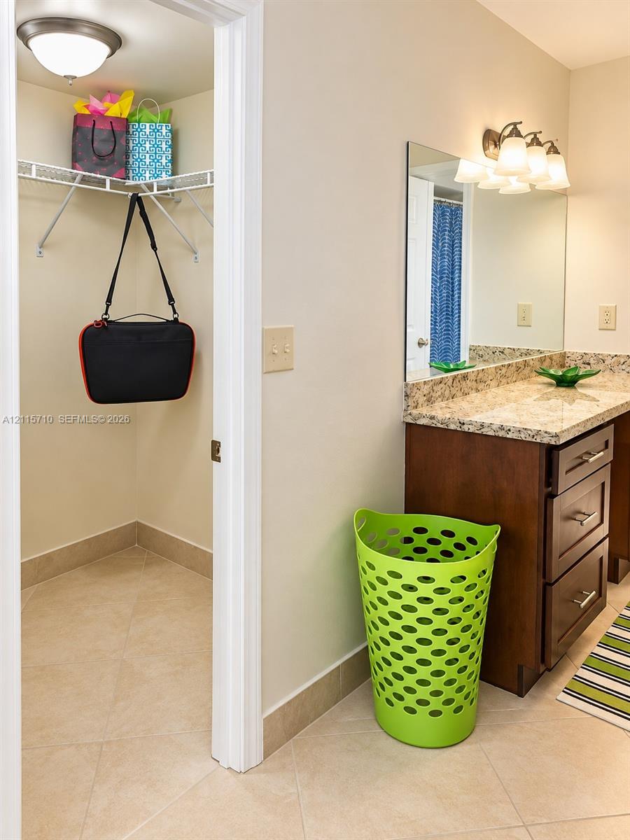 Bathroom featuring a vanity with dark wood-finish cabinetry, stone-look countertop, large vanity mirror, three-light fixture, and an internal walk-in closet with wire shelving